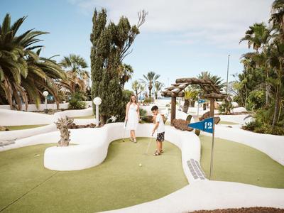 Rodina hraje minigolf na slunném, tropickém hřišti s palmami.