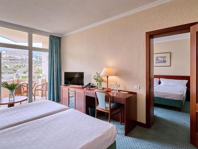 Světly hotelový pokoj s manželskou postelí, psacím stolem a výhledem na balkon a horskou krajinu.