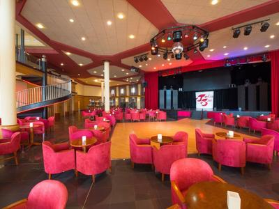 Grande salle d'événement avec fauteuils rouges et scène, éclairée par des lumières au plafond.