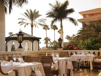 Terrasse avec tables dressées, palmiers et un kiosque dans un complexe hôtelier tropical.
