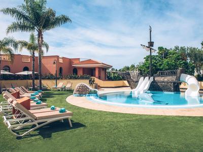 Espace piscine dans un jardin d'hôtel avec toboggans, chaises longues et palmiers.