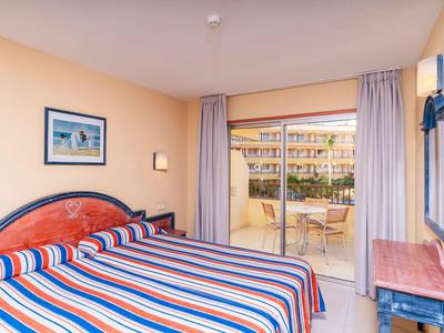 Hotelzimmer mit Doppelbett, buntem gestreiftem Bettbezug, Balkon und Blick auf umliegende Gebäude.