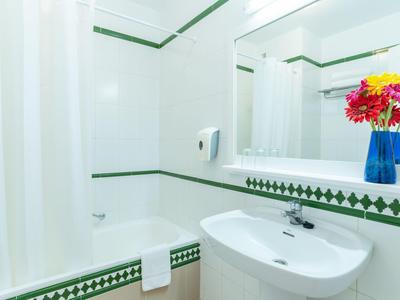 Weißes Badezimmer mit grünen Fliesen, großer Spiegel, Waschbecken und blauem Blumenvasenstrauß.