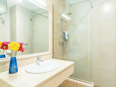 Helles Badezimmer mit weißem Waschbecken, großem Spiegel, Glasdusche und blauen Vase mit bunten Blumen.