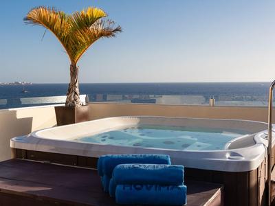 Whirlpool auf Terrasse mit blauer Decke, Palme und Meerblick bei klarem Himmel.