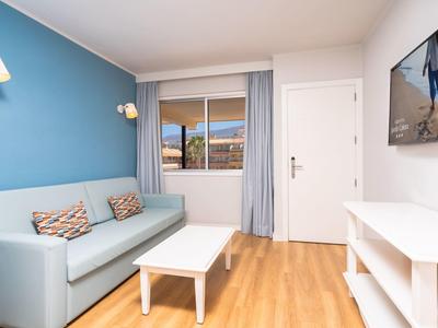 Helles Wohnzimmer mit blauer Wand, grauem Sofa, weißem Tisch und TV an der Wand, Holzboden.