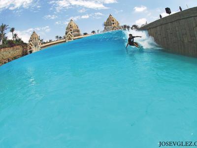 Persona surfeando una ola en una gran piscina de parque acuático con clima soleado.