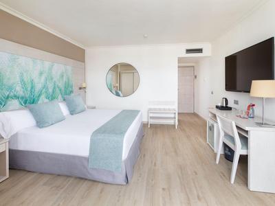 Jasny pokój hotelowy z podwójnym łóżkiem, biurkiem i okrągłym lustrem na ścianie.