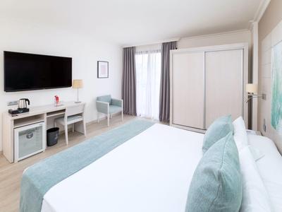 Jasny pokój hotelowy z dużym łóżkiem, telewizorem, biurkiem i małą lodówką.