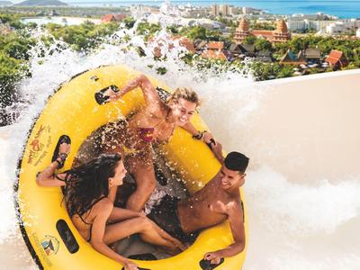 Drie mensen in een geel vlot op een golf in een waterpark met stadszicht op de achtergrond.