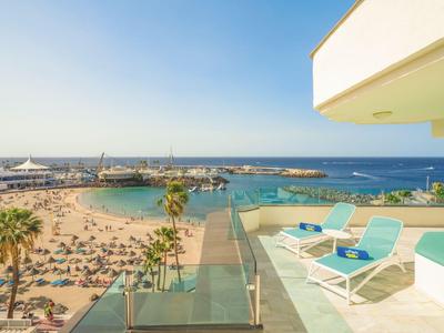 Vista desde una terraza de hotel con tumbonas hacia una playa de arena concurrida y el mar azul.