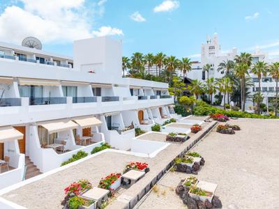 Complejo hotelero moderno con balcones blancos, palmeras y plantas en flor bajo un cielo azul.