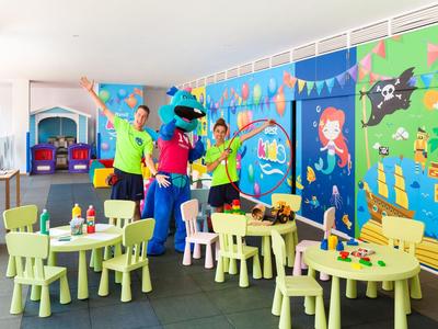 Colorful playroom with small tables and chairs, vibrant wall mural, and people celebrating.
