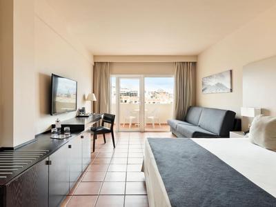 Bright hotel room with bed, sofa, wardrobe, TV, and balcony with view.