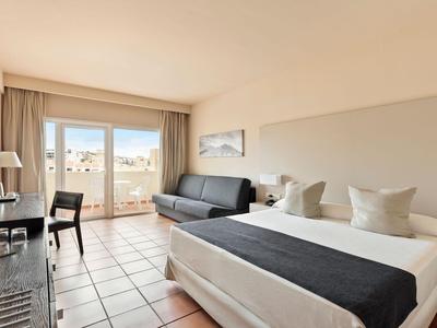 Bright hotel room with double bed, sofa, and balcony with a view.