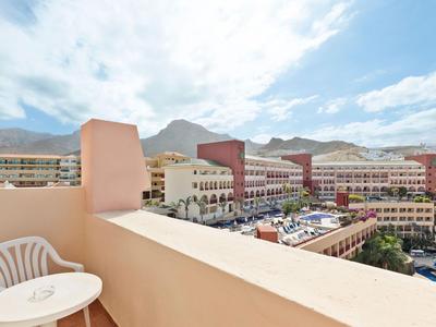 View from a balcony with chair and table overlooking hotel buildings and pool with mountain backdrop.