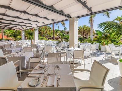 Terrazza ristorante esterna coperta con sedie bianche e tavoli apparecchiati in un ambiente tropicale.