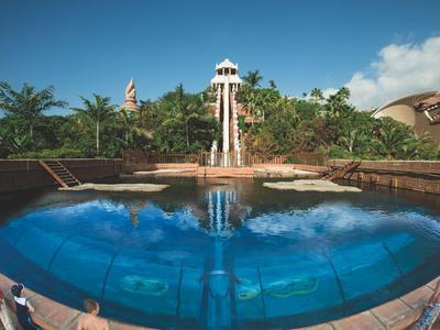 Grande piscina blu davanti a un giardino tropicale con scivolo d'acqua e lettini.