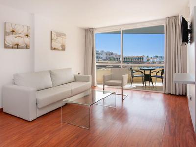 Jasny salon z białą sofą, szklanym stołem i balkonem z widokiem na morze.