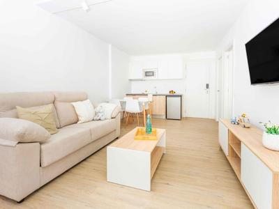 Jasny, minimalistyczny apartament hotelowy z sofą, stołem, aneksem kuchennym i telewizorem na ścianie.