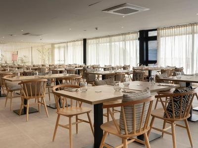 Jasna, nowoczesna restauracja hotelowa z drewnianymi krzesłami i stołami, dużymi oknami i zasłonami.