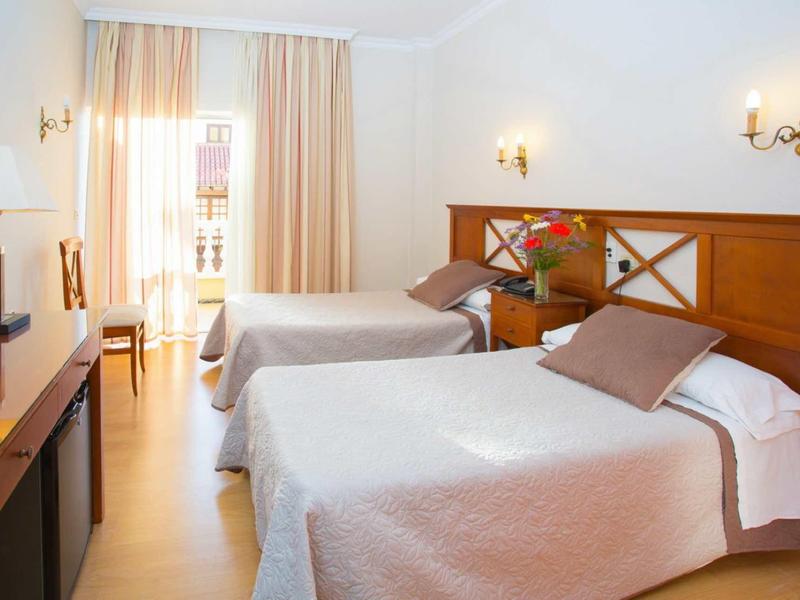 Helles Hotelzimmer mit zwei Einzelbetten, Holzmöbeln, hellen Vorhängen und warmem Licht.