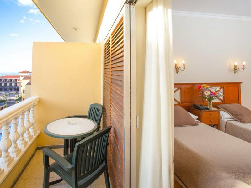 Balkon mit rundem Tisch und zwei Stühlen neben einem hellen Schlafzimmer mit zwei Betten.