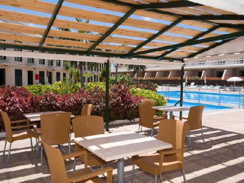 Outdoor-Terrasse mit Tischen und Stühlen unter Holzüberdachung am Pool bei Sonnenschein.