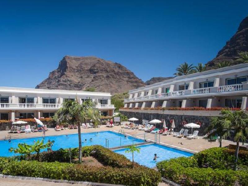 Hotel mit großem Außenpool, Liegen, Palmen und Bergen im Hintergrund bei blauem Himmel.