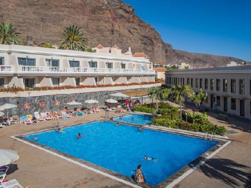 Großer blauer Pool mit Liegestühlen, Gebäuden im Hintergrund und Bergen unter klarem Himmel.