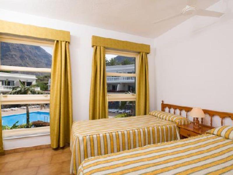 Helles Schlafzimmer mit zwei Einzelbetten, gelben Vorhängen und Blick auf Pool und Berge.