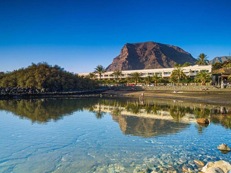 Klares Wasser spiegelt felsigen Berg und Häuser am grünen Ufer unter blauem Himmel wider.