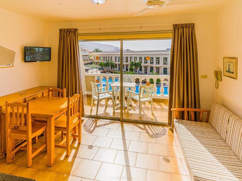 Helles Wohnzimmer mit Holzmöbeln, Fliesenboden, Sofa und Balkon mit Blick auf den Pool.