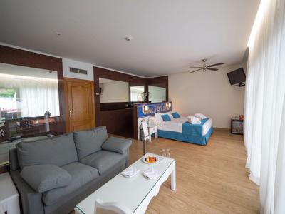 Chambre d'hôtel lumineuse avec canapé gris, lit double, sol en bois et grandes fenêtres avec rideaux blancs.