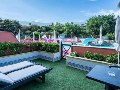 Terrasse d'hôtel avec chaises longues, table et vue sur piscine et palmiers sous ciel clair.