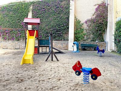 Aire de jeux pour enfants avec toboggan et jouets à ressort sur sable devant un mur d'hôtel avec plantes grimpantes.