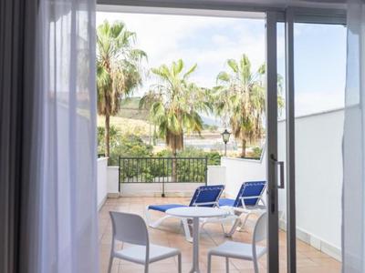Vue depuis une chambre sur une terrasse avec des chaises blanches et des transats bleus, entourée de palmiers.