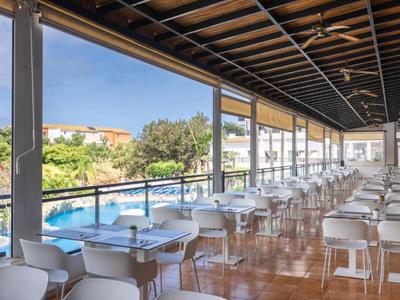 Espace de restauration ouvert avec chaises et tables blanches près d'une piscine, couvert par des poutres en bois.