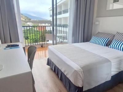 Chambre d'hôtel moderne avec lit double, bureau et balcon donnant sur les bâtiments environnants.