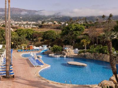 Grande piscine extérieure avec transats bleus et palmiers, entourée de verdure avec montagnes en arrière-plan.
