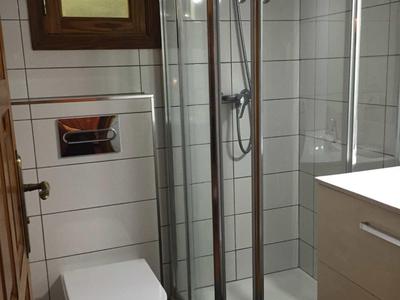 Kleines Bad mit weiß gekachelter Dusche, Toilette, Holzfenster und weißem Waschbeckenunterschrank.
