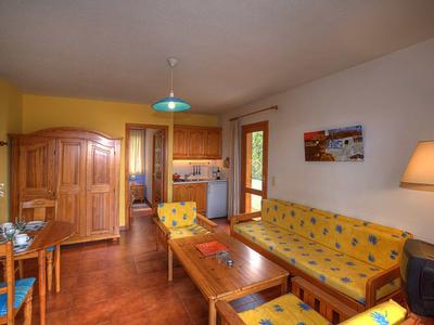 Gemütliches Wohnzimmer mit gelber Polstermöbelgarnitur, Holztisch, Schrank und offener Küche.