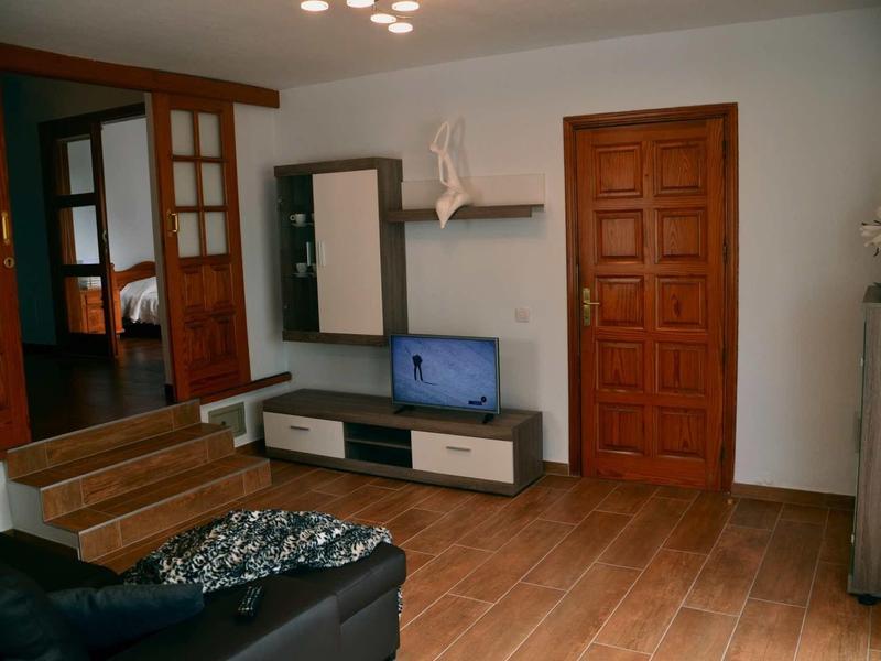 Wohnzimmer mit Holzboden, Holztür, TV auf modernem weißen Lowboard und dunklem Sofa im Vordergrund.