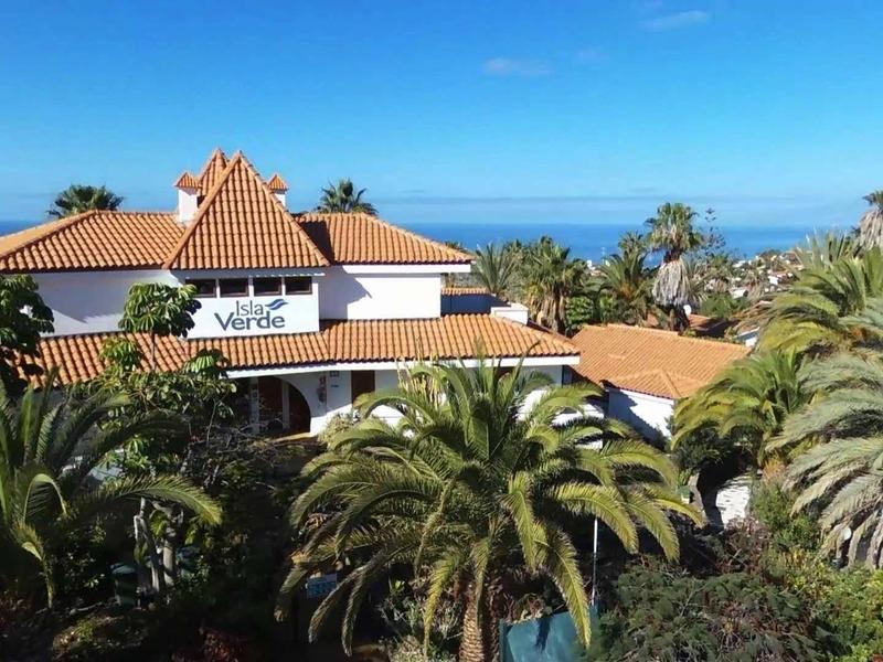 Ein Haus mit rotem Ziegeldach, umgeben von Palmen und grünem Pflanzen, blauer Himmel im Hintergrund.