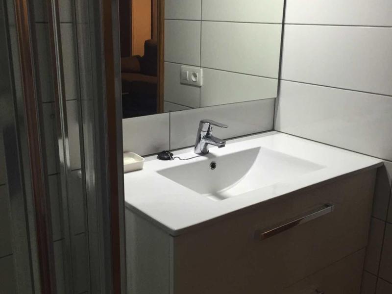 Kleines Badezimmer mit weißem Waschbecken, braunem Waschtischunterschrank und großen weißen Wandfliesen.