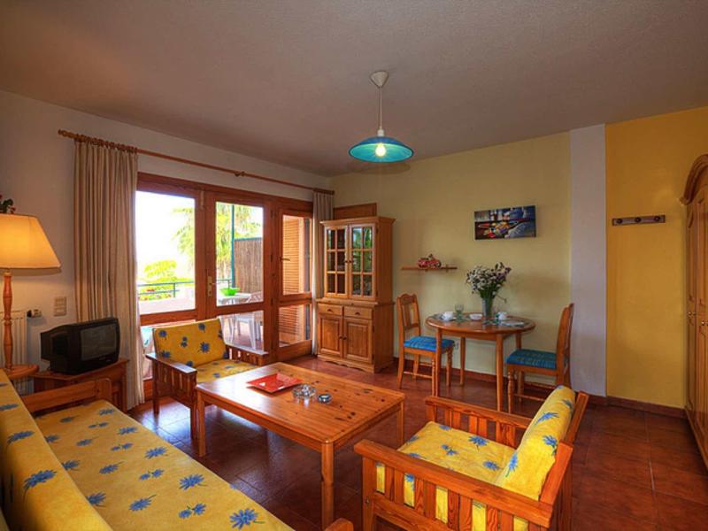 Gemütliches Wohnzimmer mit Holzmöbeln, bunten Polstern, Teppichboden und großem Fenster mit Vorhängen.