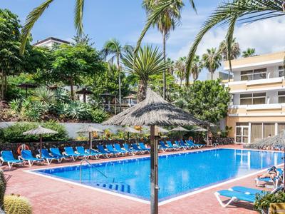 Piscine d'hôtel avec chaises longues bleues, palmiers et parasols en paille sous un ciel clair.