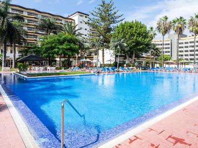 Grande piscine d'hôtel bleue avec chaises longues blanches et palmiers autour par temps ensoleillé.