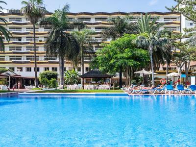 Grande piscine bleue devant un hôtel à plusieurs étages avec des palmiers et des chaises longues.