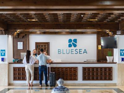 Une réception d'hôtel moderne avec trois personnes et un grand logo BLUESEA sur le mur.